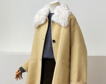 May include: A beige coat featuring a white faux fur collar, long sleeves with folded cuffs, and a single dark button closure. The coat is layered over a dark grey sweater and a dark brown skirt, displayed on a mannequin.
