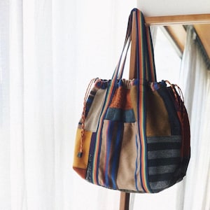 May include: A patchwork tote bag constructed from various fabric textures and colors, including mustard yellow, brown, and grey stripes. The bag features long, vibrant rainbow-striped shoulder straps and orange drawstring cords on both sides.