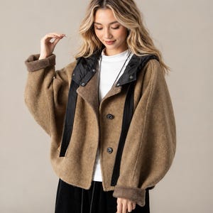 May include: A tan, textured, loose-fitting jacket with a black hood and black vertical trim along the front opening. The jacket features two dark buttons, wide sleeves with folded cuffs, and is worn over a white top and black skirt.