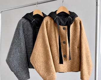 May include: Two cropped jackets hanging on wooden hangers. One jacket is tan and the other is charcoal grey. Both feature a black hood with black drawstrings and a front button closure with three dark buttons. The fabric has a fuzzy, textured finish.