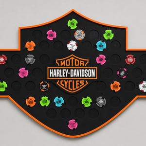 Puede incluir: Un tablero de exhibición de pared negro con forma de escudo de Harley-Davidson y borde naranja. El tablero presenta el texto MOTOR HARLEY-DAVIDSON CYCLES en el centro e incluye múltiples muescas circulares con fichas de colores.