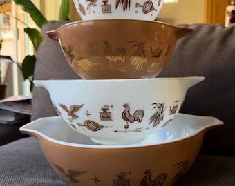 May include: A set of four nesting mixing bowls with handles, alternating between white and brown glass. Each bowl features a repeating brown graphic print of an eagle, a rooster, a coffee grinder, corn, and a kettle, arranged in a horizontal pattern.