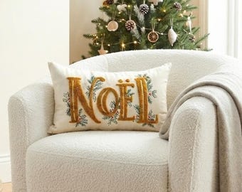 May include: A rectangular cream-colored throw pillow featuring the word NOEL embroidered in a textured gold thread. The lettering is surrounded by an intricate stitched pattern of green pine needles and small red berries, creating a festive holiday design.