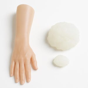 May include: A light-toned mannequin hand and forearm model positioned next to two irregular, translucent, off-white textured spheres of varying sizes. The mannequin features five fingers with defined nails and a smooth, matte skin-like finish.
