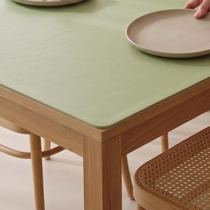May include: A rectangular dining table with a light green surface and wooden legs, accompanied by four chairs featuring woven cane backrests. The table is set with four round beige plates and folded beige fabric napkins at each place setting.