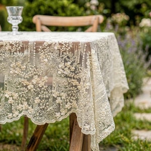 May include: A cream-coloured lace tablecloth featuring an intricate floral pattern with embroidered flowers and small white beads. The fabric is sheer and delicate, with a scalloped edge that drapes over the sides of a wooden table.