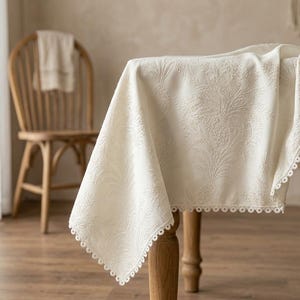 May include: An off-white tablecloth featuring an intricate embossed floral pattern across the fabric surface. The edges of the rectangular cloth are finished with a delicate, scalloped crochet trim that hangs over the side of a wooden table.