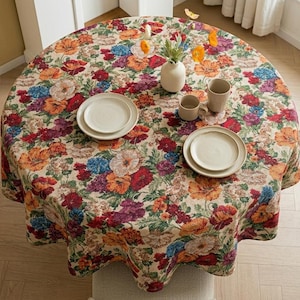 May include: A round tablecloth featuring a dense, colourful floral pattern with red, orange, purple, and blue blossoms on a cream background. The table is set with two white ceramic plates, two matching mugs, and a white vase containing orange flowers.