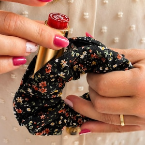 May include: A black fabric scrunchie featuring a small floral pattern in shades of red, white, and green. A gold-toned lipstick tube with a red cap labeled 156 MAT is tucked into the center of the scrunchie, showing the product branding.