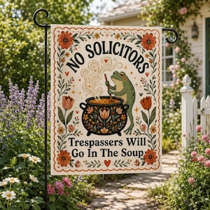 May include: A rectangular garden flag featuring a green frog stirring a black cauldron with floral patterns. The text reads NO SOLICITORS and Trespassers Will Go In The Soup. The design is framed by orange flowers and green leaves on a cream background.