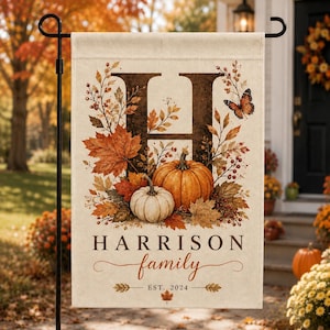May include: A rectangular garden flag featuring a large dark brown letter H surrounded by autumn leaves, berries, a butterfly, and two pumpkins. The text reads HARRISON family EST. 2024. The flag is displayed on a black metal stand outdoors.