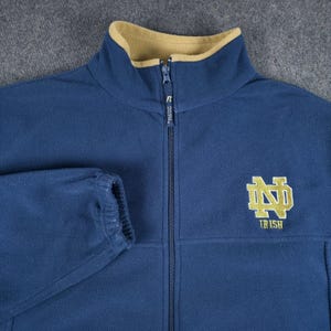 Puede incluir: Una chaqueta de forro polar azul marino con cremallera, cuello interior color canela y un logotipo de Notre Dame bordado en dorado en el pecho. El tirador de la cremallera lleva la letra R. La chaqueta tiene mangas largas con puños elásticos.