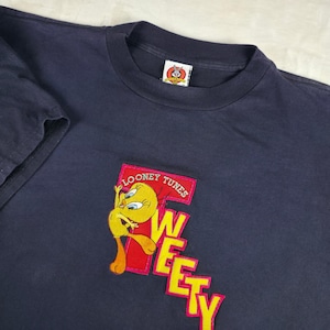 May include: A dark navy blue short-sleeve t-shirt featuring a large embroidered graphic of the yellow Tweety Bird character. The design includes the red text Looney Tunes and the yellow text Tweety. A Looney Tunes brand tag is at the neckline.