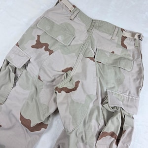 May include: Cargo pants featuring a desert camouflage pattern in shades of tan, beige, and brown. The pants include two rear pockets with flaps, side cargo pockets, belt loops, and adjustable side waist tabs with metal buckles for a secure fit.