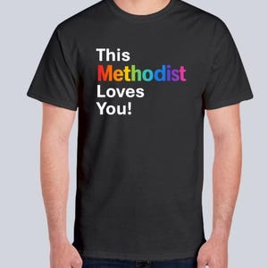 May include: A black short-sleeved t-shirt featuring the white text "This" and "Loves You!" with the word "Methodist" printed in a multi-coloured rainbow gradient. The shirt has a crew neckline and a standard fit, displaying clear, bold typography.