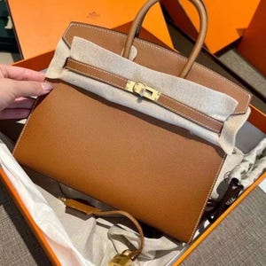 May include: A tan leather handbag with a textured surface, featuring two top handles, a gold-tone metal turn-lock closure, and a matching leather strap with a small gold-tone padlock attached. The bag is partially covered with protective white paper.