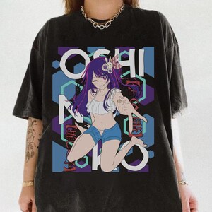 May include: A black short-sleeve t-shirt featuring a graphic print of an anime character with purple hair, a white ruffled top, and blue denim shorts. The background includes blue and purple hexagonal shapes and the text OSHI NO KO in white.