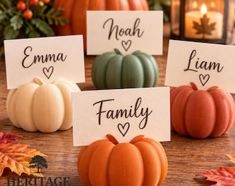 May include: Four small, decorative pumpkin-shaped place card holders in cream, sage green, burnt orange, and terracotta. Each holds a white rectangular card printed with the names Emma, Noah, Liam, and the word Family, each with a heart icon.
