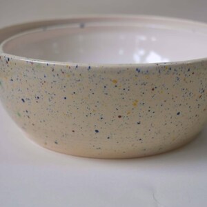 Peut inclure: Un bol en céramique avec un extérieur de couleur crème présentant un motif moucheté de points bleus, jaunes et verts. L'intérieur du bol est lisse et blanc, avec un bord arrondi et une large base circulaire qui s'affine vers le bas.