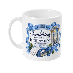 May include: A white ceramic mug featuring a blue ribbon graphic with the text The Order of Women Freemasons, Congratulations on becoming an Entered Apprentice, and The First Degree. It includes illustrations of a white glove, a trowel, and a book.