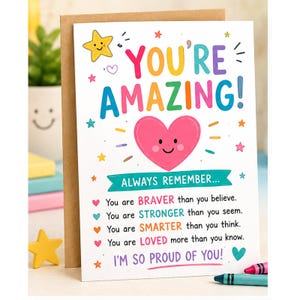 Può includere: Un biglietto d'auguri bianco con il testo colorato YOU'RE AMAZING! sopra un cuore rosa con una faccia sorridente. Sotto, un nastro turchese riporta ALWAYS REMEMBER... seguito da quattro righe di testo in vari colori e la frase I'M SO PROUD OF YOU!