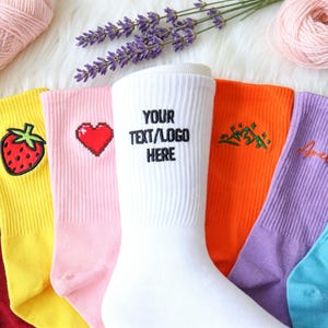May include: A collection of crew socks in various colours including red, yellow, pink, white, orange, purple, and blue. Each sock features unique embroidery, such as a letter R, a strawberry, a heart, a mountain, a name, and the text YOUR TEXT/LOGO HERE.