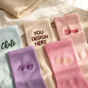 May include: Five ribbed crew socks in pastel colors including light blue, purple, cream, bright pink, and light pink. Each sock features custom embroidery with names like Chole, Ashley, Emma, and Nahia, or the text YOU DESIGN HERE.