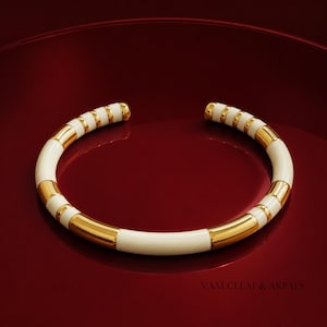 May include: A gold and cream-colored open cuff bracelet featuring alternating smooth metallic gold segments and smooth cream-colored segments. The bracelet has a circular shape with a small opening at the top and the text VAM CLEAF & ARPALS.