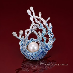 Puede incluir: Un broche con forma de ramas de coral que presenta un degradado de piedras preciosas azules y blancas con una perla blanca central. Incluye el texto VAM CLEAF & ARPALS en la parte inferior derecha sobre un fondo rojo con reflejo.