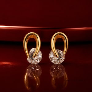 Puede incluir: Un par de pendientes dorados, cada uno con un bucle de metal liso y curvado que rodea parcialmente una piedra preciosa redonda, transparente y facetada. Los pendientes se muestran sobre una superficie oscura y reflectante, creando un reflejo.