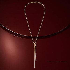 Puede incluir: Un collar de tono dorado con una cadena fina y lisa que forma un diseño de nudo central. El collar incluye dos extremos de cadena que cuelgan hacia abajo desde el nudo y un pequeño cierre de mosquetón en la parte superior con una cadena ajustable.