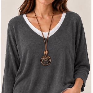 May include: A charcoal grey V-neck top layered over a white shirt, paired with a long brown leather cord necklace featuring two wooden beads and three concentric circular metal pendants with a detailed cutout design in the centre.