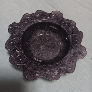 May include: A circular black dish with a glittery finish and a scalloped, decorative edge. The center is smooth and recessed, while the wide rim features a textured, embossed pattern that follows the irregular, wavy shape of the outer border.