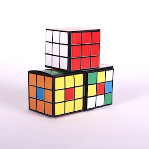 Puede incluir: Tres cajas de forma cúbica de colores apiladas una encima de la otra. La caja superior es roja y blanca, la caja del medio es verde y blanca, y la caja inferior es naranja, amarilla, azul y blanca.