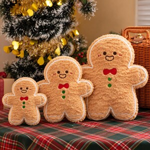 May include: Three plush gingerbread man toys in varying sizes, each featuring a textured tan fabric, white icing-style trim, a red bow tie, two green buttons, and a smiling face with rosy cheeks, arranged in a row on a red and green plaid surface.
