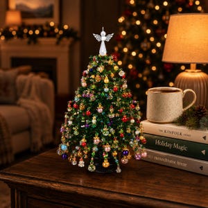 May include: A miniature green beaded Christmas tree decorated with various colorful hanging ornaments and topped with a white angel figure. The tree sits on a dark base, positioned next to a stack of three books and a ceramic mug.