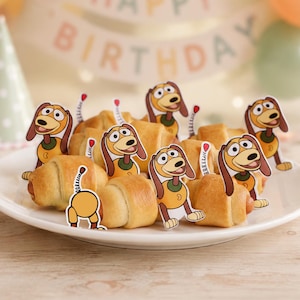 May include: A white plate filled with golden-brown crescent rolls, each decorated with paper toppers featuring a cartoon dog character with a long spring-like body, brown ears, and a yellow torso, arranged to look like a dachshund toy.