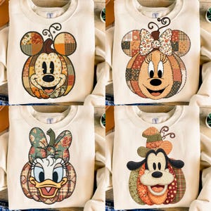 May include: Four cream-colored sweatshirts featuring graphic prints of cartoon characters shaped like pumpkins. The designs include a mouse with round ears, a mouse with a bow, a duck with a bow, and a dog with a hat, all in plaid patterns.