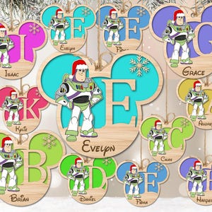 May include: A collection of wooden Mickey Mouse-shaped Christmas ornaments featuring Buzz Lightyear wearing a Santa hat. Each ornament displays a large letter, a personalized name, and a snowflake cutout in various colors like pink, blue, and green.