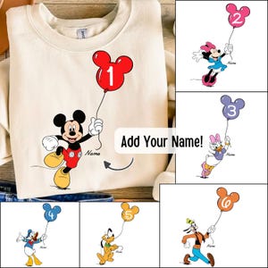 Puede incluir: Una sudadera de cuello redondo color crema con un gráfico de Mickey Mouse sosteniendo un globo rojo con el número uno. El diseño incluye el texto Name y Add Your Name. Paneles adicionales muestran a Minnie, Daisy, Donald, Pluto y Goofy.