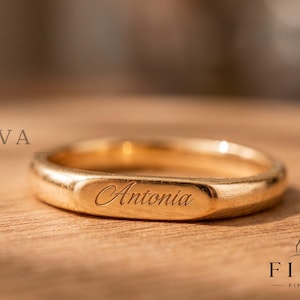 May include: A polished gold band ring featuring a flat, rectangular front surface engraved with the name Antonia in a cursive script. The ring has a smooth, rounded shank and a metallic finish, with the text FIORVA FINE JEWELLERY visible.