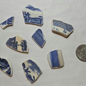 May include: Eight irregular ceramic shards featuring blue and white patterns, including floral and geometric designs, arranged alongside a standard 24.26 mm diameter coin for scale to indicate the size of the broken pottery fragments.