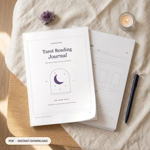 May include: A white Tarot Reading Journal with a purple crescent moon graphic inside an arch, featuring the text "Tarot Reading Journal," "Make space for intuition," and "READ / RECORD / REFLECT." A dark blue pen rests on the open pages.