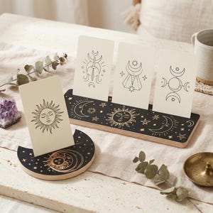 Puede incluir: Un conjunto de soportes para tarjetas de madera negra con motivos celestiales dorados que incluyen soles, lunas y estrellas. El set incluye una base rectangular con tres ranuras y una base en forma de media luna con una sola ranura.