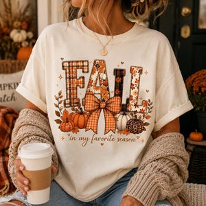 Peut inclure: Un t-shirt à manches courtes de couleur crème avec un motif représentant le mot FALL en grandes lettres, décoré de motifs à carreaux, floraux et léopard. Sous le texte se trouve un grand nœud à carreaux orange, des citrouilles et le texte in my favorite season.