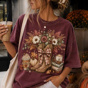 Peut inclure: Un t-shirt à manches courtes bordeaux avec une impression graphique représentant deux bottes de cow-boy avec un nœud à carreaux, une citrouille, une tasse de café, une bougie et un arrangement floral, porté avec une casquette bordeaux.