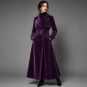 Könnte beinhalten: Ein langer Trenchcoat aus tiefviolettem Samt mit zweireihiger Knopfleiste, dunklen Knöpfen, einem passenden Stoffgürtel mit rechteckiger Schnalle, breitem Revers und langen Ärmeln. Der Mantel reicht bis zum Boden und hat eine taillierte Passform.