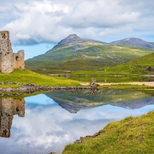 May include: A stone castle ruin stands on a grassy hill beside a calm lake, reflecting the structure and the surrounding green mountains in the water. The sky above is filled with white clouds, and the landscape features vibrant green hills.