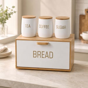 May include: A rectangular wooden bread box with a white front panel featuring the word BREAD in gold lettering and a wooden handle. Three white canisters with wooden lids labelled TEA, COFFEE, and SUGAR in gold text sit on top of the box.