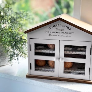 May include: A white wooden egg cabinet shaped like a house with two doors featuring wire mesh panels and small white knobs. The front is printed with the text GENERAL STORE Vintage FARMERS MARKET Groceries & Provisions. Brown eggs are visible inside.